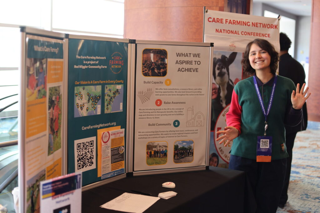 person smiling and standing in front of a Care Farming Network exhibit at a conference